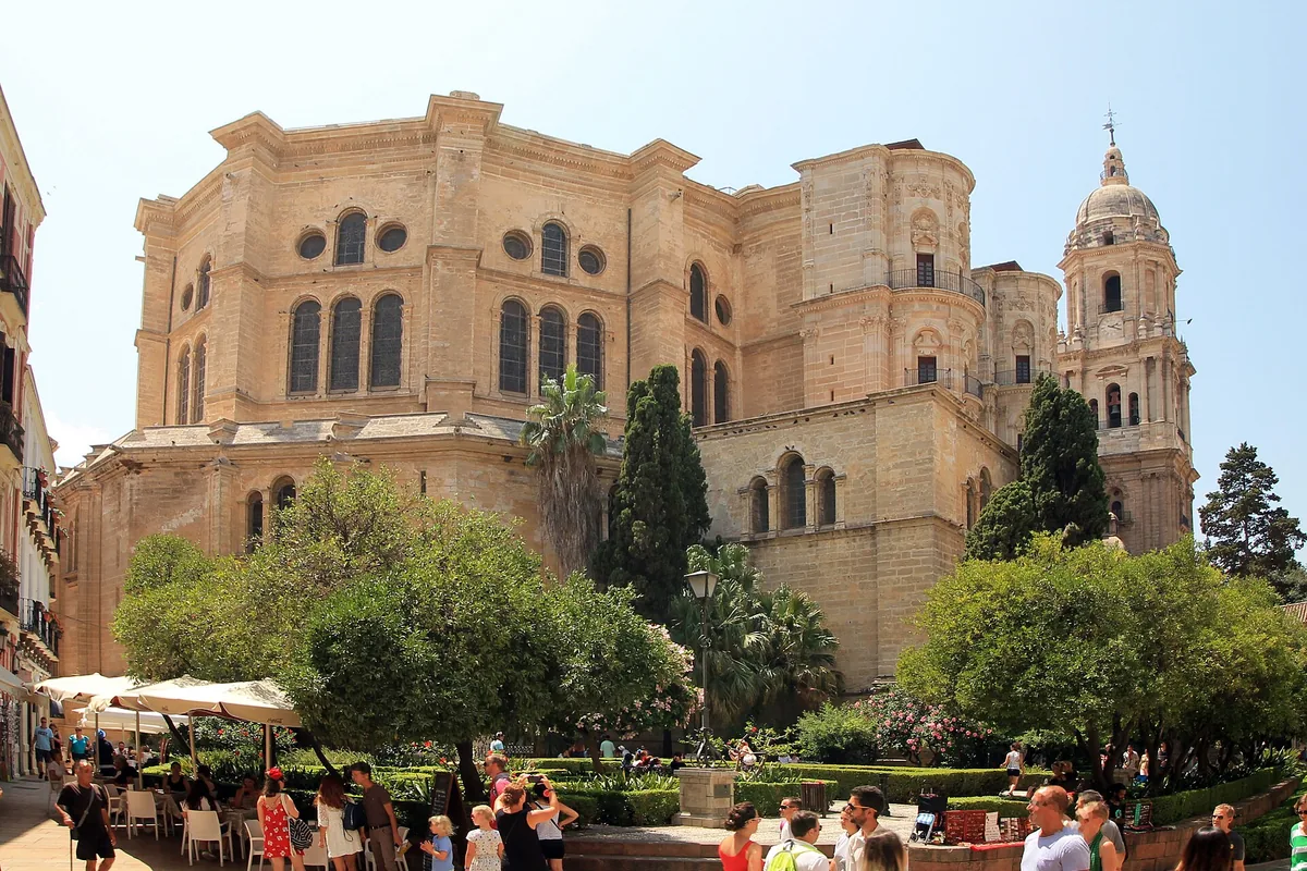 Málaga Cathedral seen from the north-east in daylight, a sandy stone Renaissance building with one finished bell tower, behind orange trees