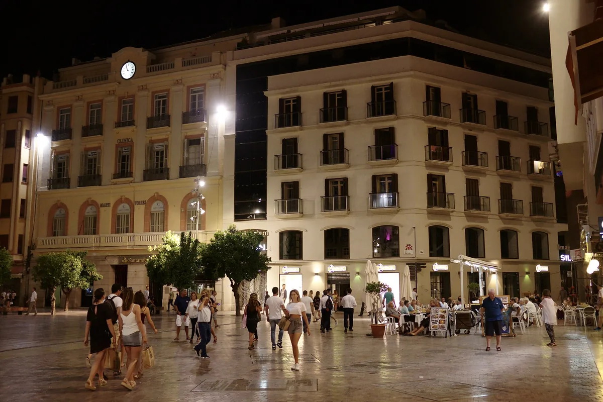 Plaza de la Constitución in Málaga at night, with pale buildings lit from below, a clock, trees and people crossing the square