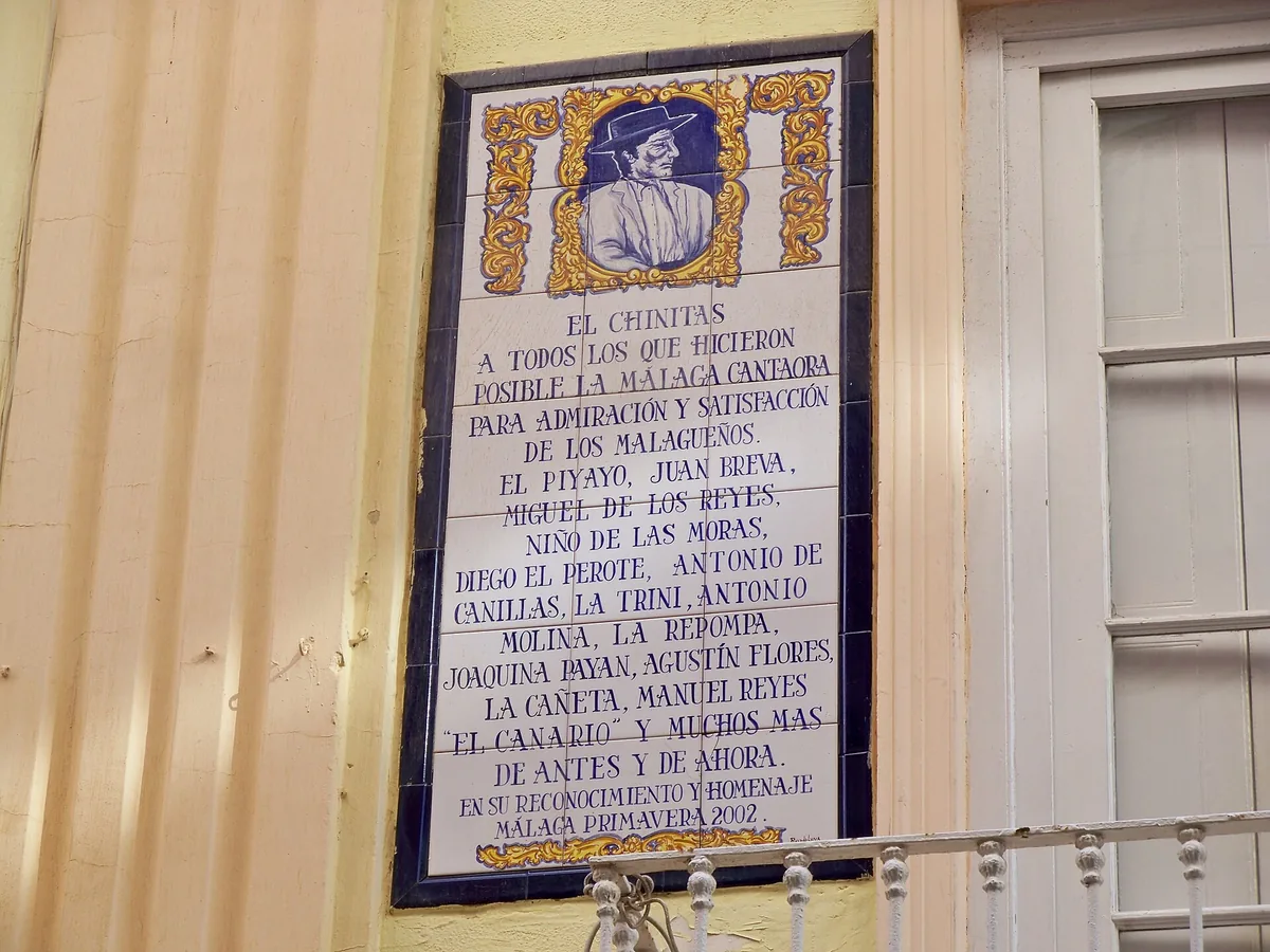 A blue and gold tile plaque headed El Chinitas listing flamenco singers of Málaga, set into a cream wall beside a window